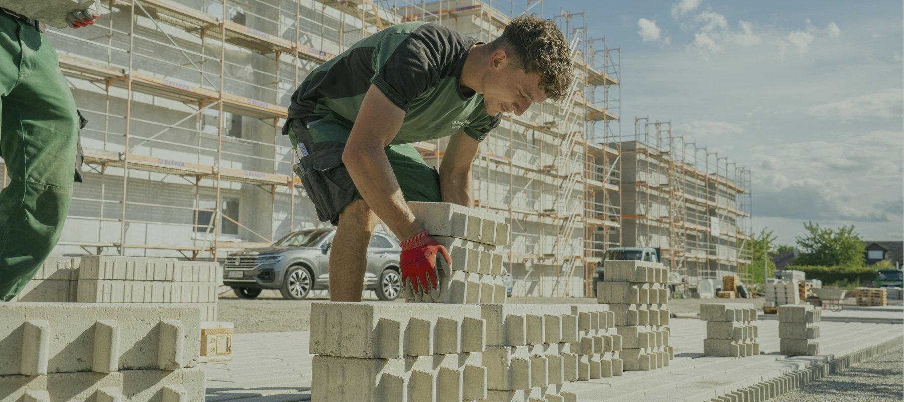 Mitarbeiter verlegt Pflastersteine auf einer Baustelle vor einem eingerüsteten Neubau