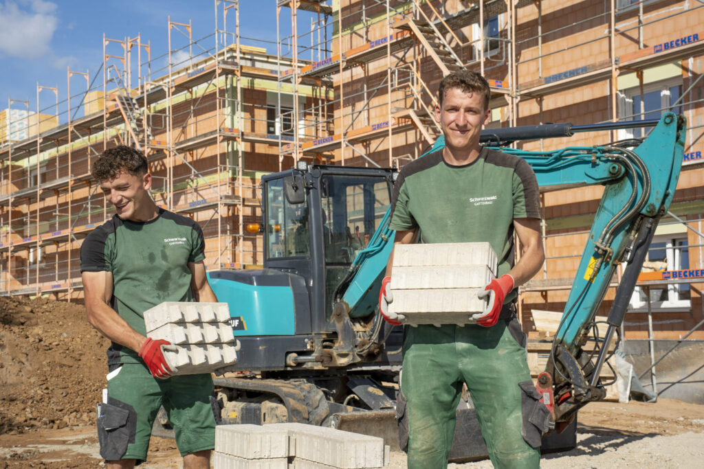 Zwei Mitarbeiter von Schwarzwald Gartenbau tragen Pflastersteine vor einem Minibagger und einem eingerüsteten Neubau.